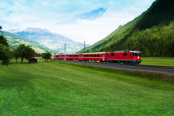 Red train crossing green valley near Alps