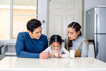 Portrait of enjoy happy love asian family father and mother with little asian girl smiling and having protein breakfast drinking and hold glasses of milk at table in kitchen.