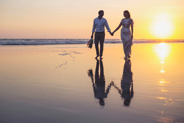 Newlywed couple holding hands while walking at beach against clear sky during sunset