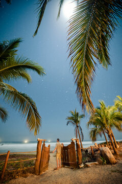 A man opens a wooden gate leading to the beach under a full moon and starry night sky in Mexico.