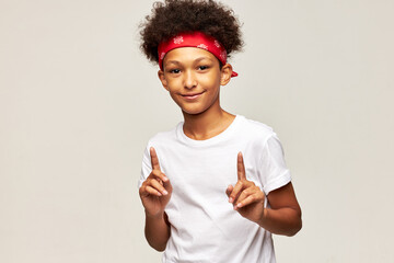 Attention, please! Studio image of happy african american teen boy pointing index fingers up posing in white sport mock-up t-shit with copy space for your advertising text, isolated on gray background - Powered by Adobe