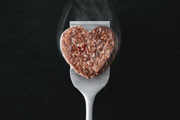 heart shaped beef burger patty on a grilled spatula. dark background place for text. valentines day celebration concept