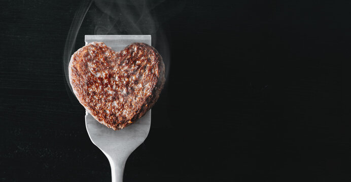 heart shaped beef burger patty on a grilled spatula. dark background place for text. valentines day celebration concept