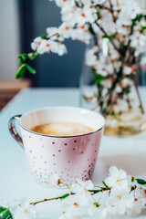 A cup of coffee in a light pink cup and spring blooming branch with flowers on the white table background. Minimalist Vertical card. Soft selective focus, copy space. Tender Spring morning mood.