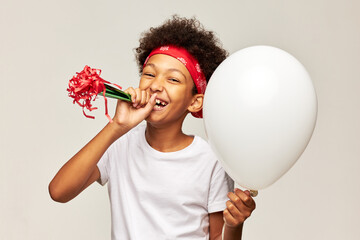 Afro kid in bandana having fun celebrating anniversary or cheering his friend laughing out loud trying to blow in horn while holding white helium balloon with copy space for your content