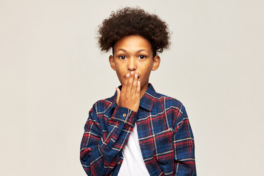 Oops. Cute african american 13-years-old boy in plaid shirt standing against gray studio background with shocked face covering opened mouth with palm, feeling guilty for making mistake