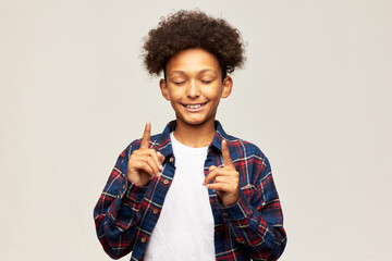 Funny picture of handsome kid explaining something, pointing index fingers up with closed eyes, trying to remember something, isolated on gray studio background. Body language, signs, gestures