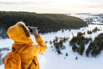 Young woman in yellow looking through binoculars at birds on snowy river against winter forest...