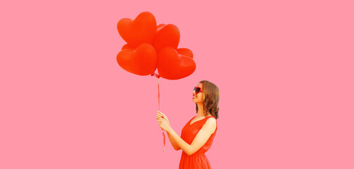 Portrait of happy smiling young woman with red heart shaped balloons having fun wearing sunglasses on pink background