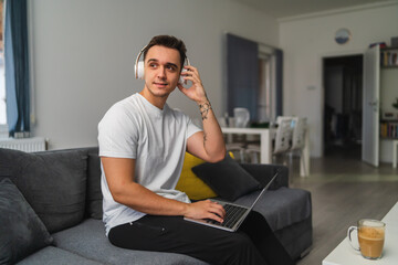 A young guy is using laptop and drinking coffee while working from home from his couch 