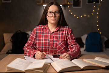 Happy clever caucasian female student in casual wear, sits at the table with books in the university dormitory, takes notes in a notebook, looking at the camera. Back to school.