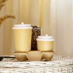 Coffee to go. Coffee composition with two disposable eco friendly cardboard cups on wicker basket in light interior. Takeaway coffee concept. Morning coffee still life. Front view, square format