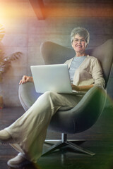 woman sitting on modern chair near the window in light cozy room at home. She is working on laptop in relaxing atmosphere