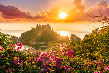 Fototapeta na wymiar sunrise or sunset view to a beautiful isle in sea from green and red flower bushes on foreground with clouds on the background of landscape