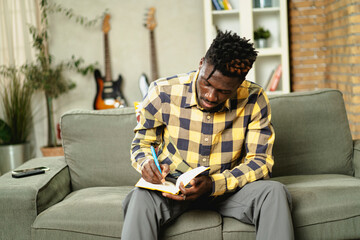Young African man writing down notes while sitting on sofa at home.