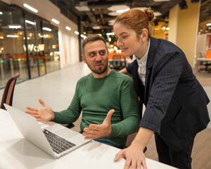 A red-haired Caucasian woman stands at the desktop of a bearded man. The boss approves the work of...