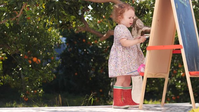 Funny baby girl wearing red rain boots paints outdoor in beautiful orange garden