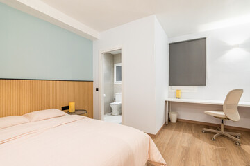 View of bedroom with large bed, work area with white wooden table and comfortable leather office chair. Small private bathroom with open door. To side is comfortable armchair with floor lamp.