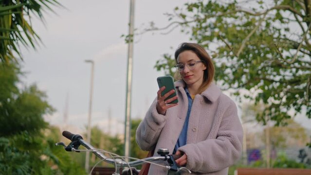 medium shot of a young hipster woman walking around an urban park with her bicycle and checking her social networks on a smartphone