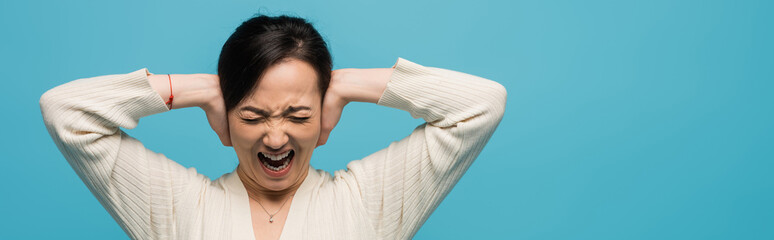 Aggressive asian woman screaming and covering ears isolated on blue, banner