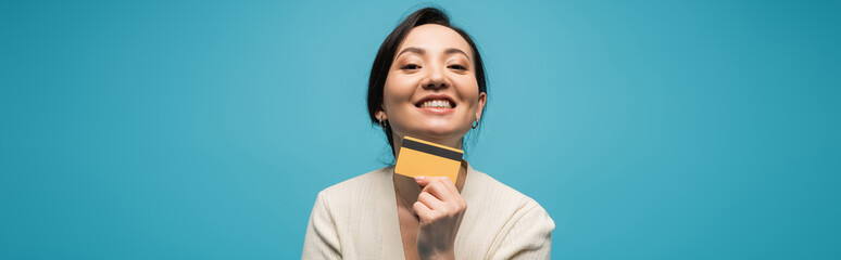 Smiling asian model holding credit card isolated on blue, banner