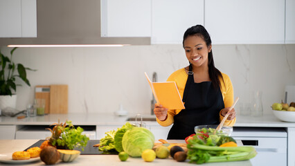 Smiling pretty millennial black female in apron making salad, reading new recipe in notebook book