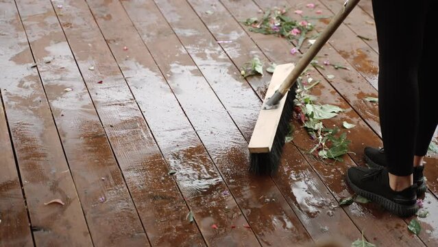 Slender woman in black leggings sweeps cut leaves and flower petals into pile with wooden brush. Garbage lies on wooden plank floor with wet puddles after rain, close-up.