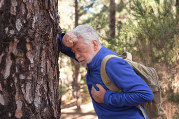 Senior bearded man with backpack feeling bad while walking in the mountain woods. Touching his...