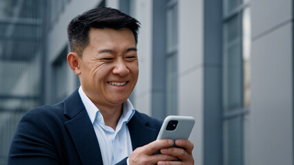Emotional smiling laughing Asian middle-aged Korean businessman man employer entrepreneur laugh looking at mobile phone chatting browsing smile watching funny video texting business in city outdoors