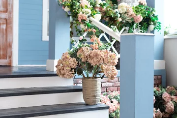 Rolgordijnen Spring design home with plants and bloom flowers hydrangea in pot on steps. House entrance staircase at home decorated for easter. Wooden porch of house with different flowers. Terrace of summer house © stock_studio