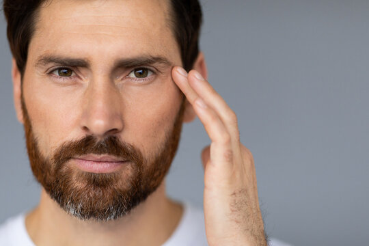 Male facial skincare. Bearded middle aged man touching face, caring for skin under eyes, grey background, copy space