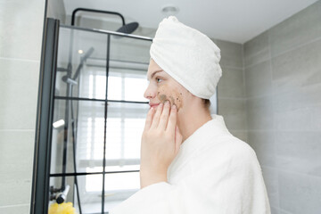 Woman in towel turban applying exfoliating cream on face in bathroom