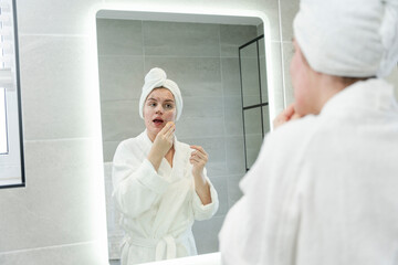 Woman in bathrobe cleaning face in front of bathroom mirror