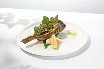 Whole roasted fish with spinach leaves on white plate. Grilled dorado on light background in summer menu. Healthy food - Roasted dorado fish. Concept menu with hard shadows.