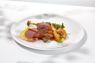 Traditional france dish - duck confit with baked apple on light background with shadows of sunlight. Roasted duck leg with fruits and gravy on white plate in summer dining Elegant roasted leg