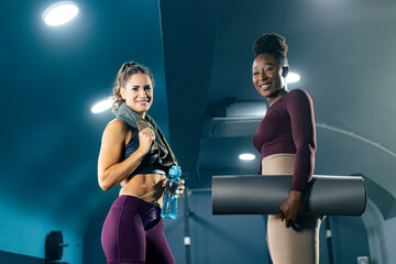 Portrait of two fit women in sportswear holding mat, bottle of water and towel in gym.