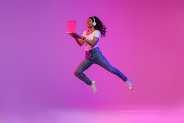 Happy Black Woman In Wireless Headphones Jumping With Laptop In Neon Light
