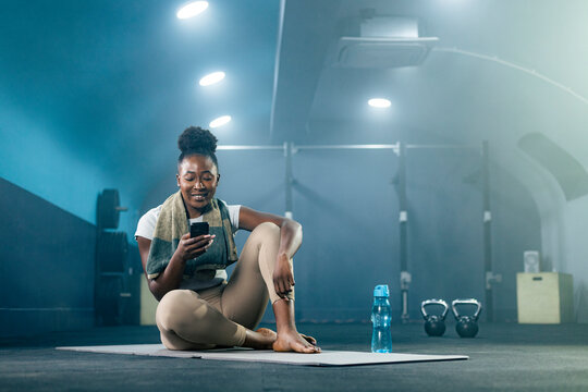 Fit African American woman in sportswear using smart phone at gym. She is sitting on yoga mat.