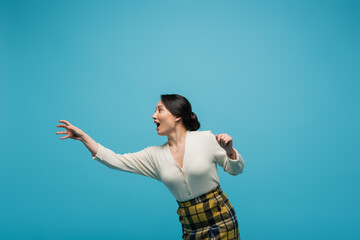 cheerful asian woman in plaid skirt smiling while gesturing isolated on blue