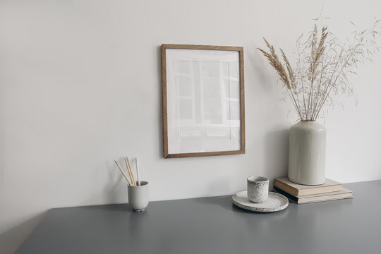 Moody wokspace still life. Pencils in ceramic holder. Vase with dry grass and cup of coffee. Blank vertical picture frame mockup hanging on white wall background. Grey table background. Home office.