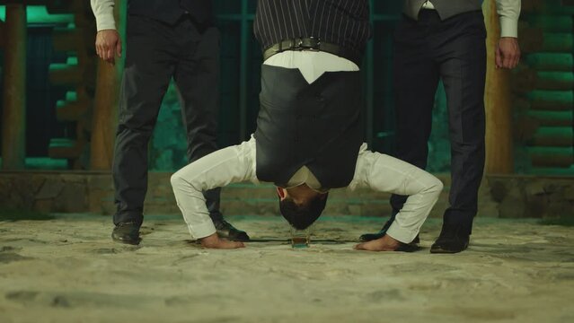 Young man flipping upside down on the floor , doing trick with glass of alcohol drink , drinking . His friends applauding cheering him . Men celebrating , having fun, laughing . Drinking without hands