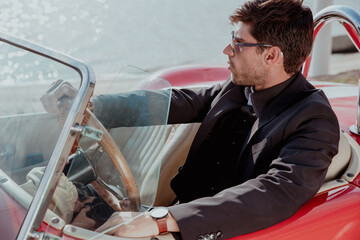 young man in convertible car looking at camera, smiling, man wears suit and tie drives red convertible car