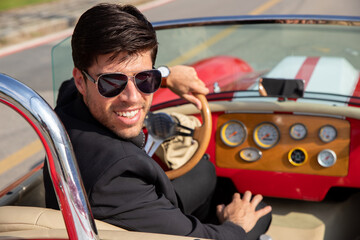young man in convertible car looking at camera, smiling, man wears suit and tie drives red convertible car
