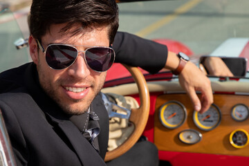 young man in convertible car looking at camera, smiling, man wears suit and tie drives red convertible car