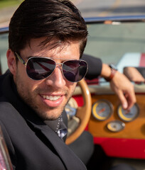 young man in convertible car looking at camera, smiling, man wears suit and tie drives red convertible car