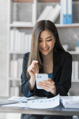 Texting message on mobile, Beautiful Asian female bookkeeper doing online accounting with laptop in private accounting office, balance sheet and stock market statistics.