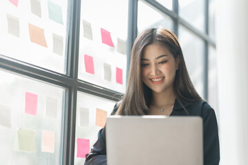 Using laptop, Pretty asia business woman bookkeeper brainstorming and working in modern office workplace with sticky notes or post it at windows.