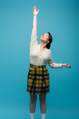 young asian woman in plaid skirt standing with outstretched hand and looking up isolated on blue