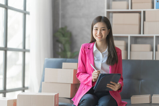 Young pretty asian business woman in suit working with product stock while sitting at sofa, preparing and checking customer address on packaging box.