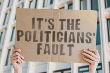 The phrase " It's politicians' fault " is on a banner in men's hands with blurred background. Bribery. Dishonesty. Fraud. Impunity. Lapses. Misrepresentation. Waste. Abusive. Betrayal. Deception. Defi